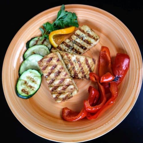 3 slices of grilled tofu on orange plat with grilled red peppers and zucchini