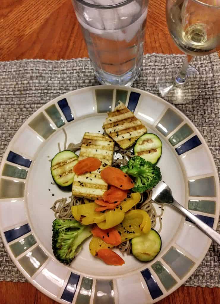 grilled tofu with carrots, broccoli, zucchini, and yellow peppers over buckwheat noodles on plate with glasses of water and wine.