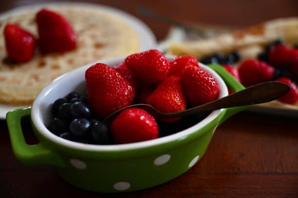 strawberries and blueberries in small bowl with spoon with crepes in background