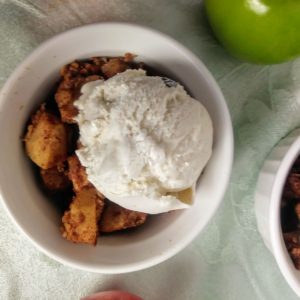 white dish of apple crisp topped with vegan vanilla ice cream next to green apple