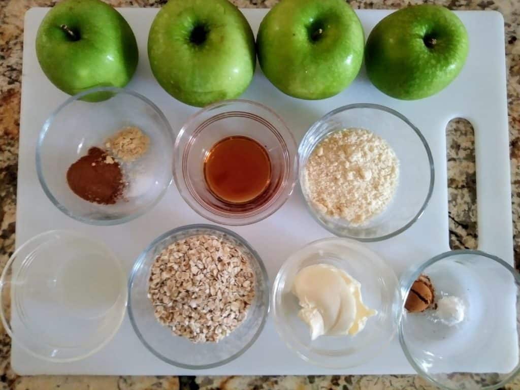 4 green apples with bowls of other ingredients like oats, almond flour, and cinnamon on white cutting board