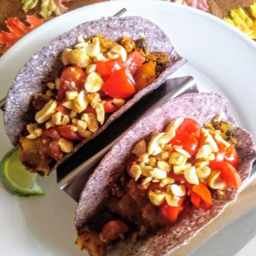2 pumpkin and black bean tacos in blue taco shells topped with chopped peanuts and a lime wedge on plate
