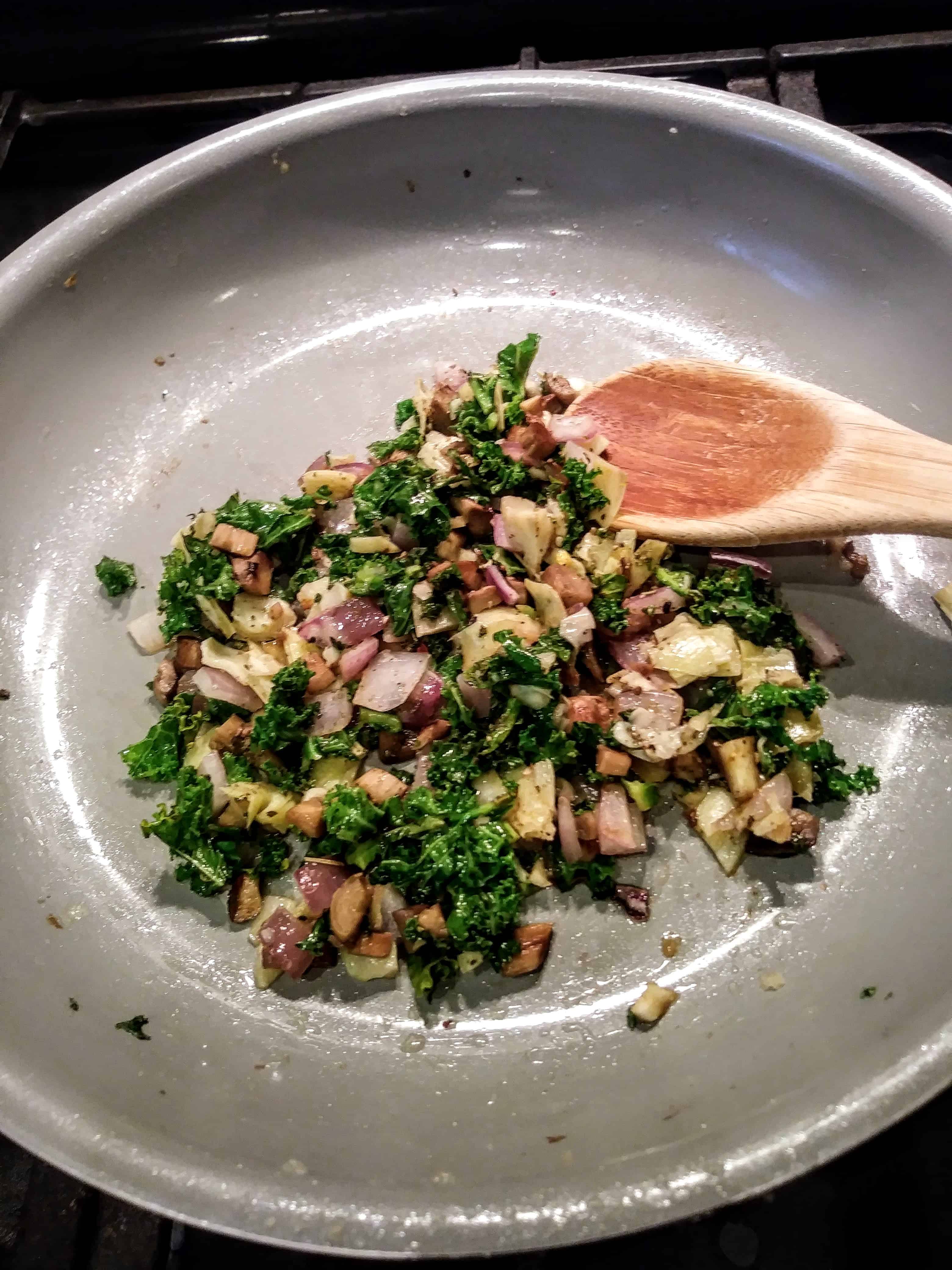 Onions, kale, artichokes, and mushroom stems cooking in pan.