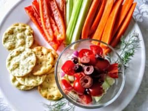 vegan hummus layered dip on plate with crackers, red pepper strips, celery sticks, and carrot sticks.