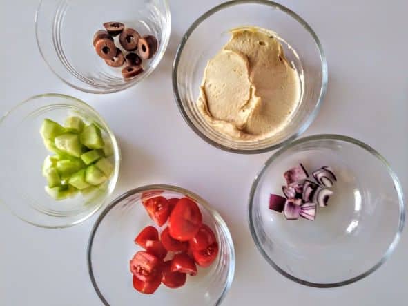 hummus, chopped tomatoes, cucumber, sliced olives, and chopped red onions in glass bowls