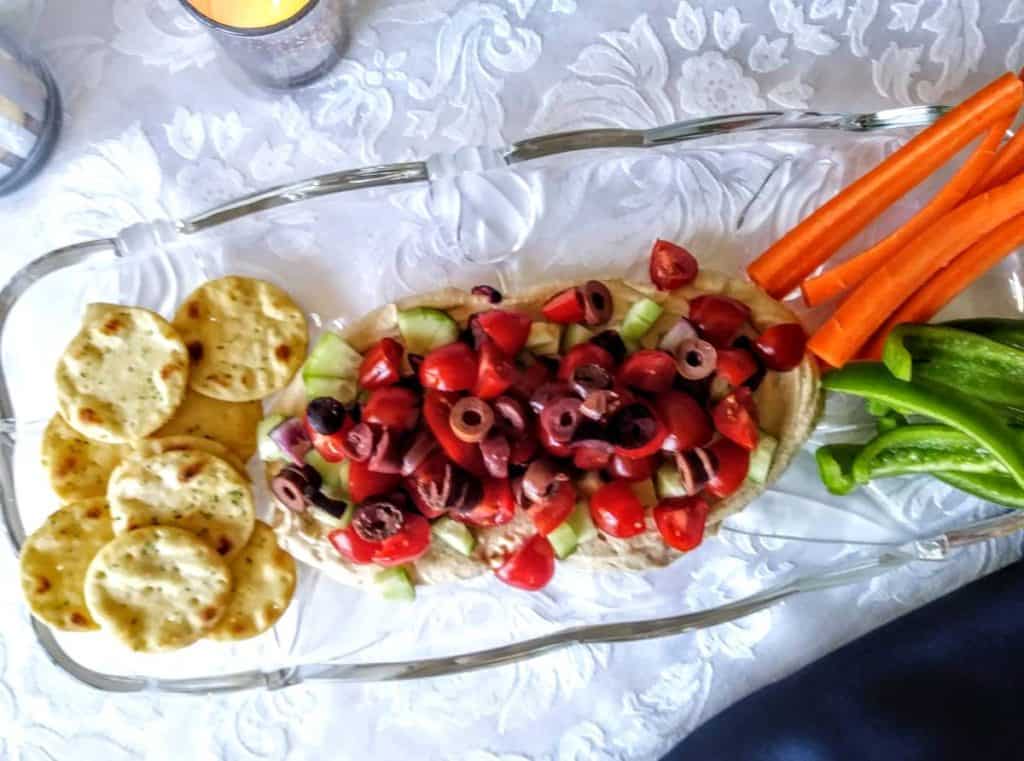 glass platter with vegan hummus layered dip, pita crackers, green peppers, and carrot sticks