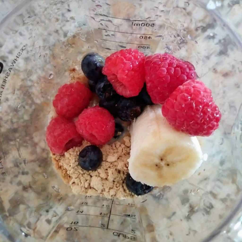 peanut butter smoothie ingredients in blender cup with raspberries, blueberries, and bananas