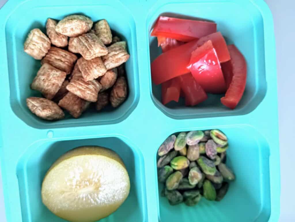 Container with Puffins cereal, red peppers, yellow kiwi half, and pistachios.